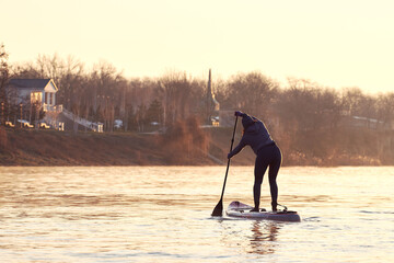 Rear view of middle-aged woman stand up paddle boards on a Danube river at sunrise on a clear winter morning