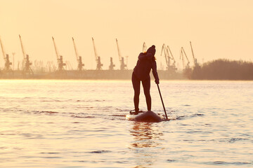 Silhouette of middle-aged woman stand up paddle boards on a Danube river at winter sunrise against...