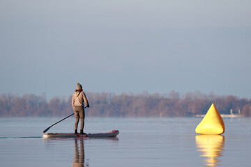 Woman on stand up paddle board at dawn in a winter river near yellow buoy