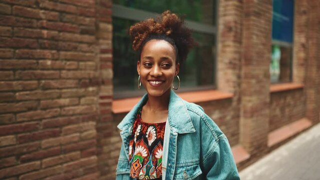 Close-up African girl with ponytail wearing denim jacket, in crop top with national pattern walking smiling down the street against modern buildings background. Slow motion