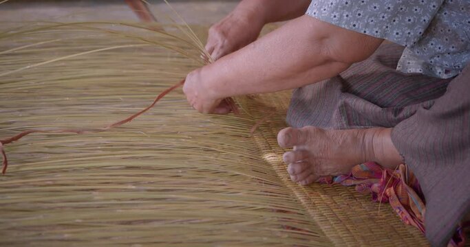 Cut in shot of an Asian female farmer who is an elderly native basketry artisan weave mat from bamboo that are cut into strips for weaving folk crafts in rural areas of Thailand, southeast Asia.