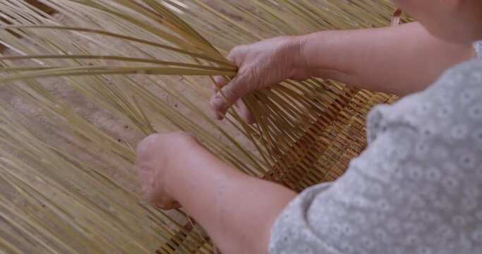 Cut in shot of an Asian female farmer who is an elderly native basketry artisan weave mat from bamboo that are cut into strips for weaving folk crafts in rural areas of Thailand, southeast Asia.