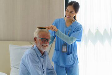 Young Asian woman nurse helping brushing hair to disabled elderly man in bed at retirement home. Millennial caregiver assisting handicapped senior patient, taking care of older male indoors