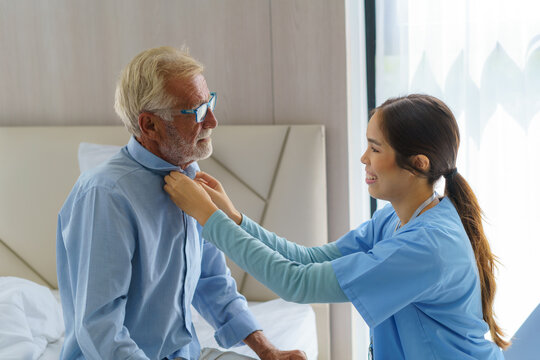 Young Asian Woman Nurse Helping Get Dress To Disabled Elderly Man In Bed At Retirement Home. Millennial Caregiver Assisting Handicapped Senior Patient, Taking Care Of Older Male Indoors