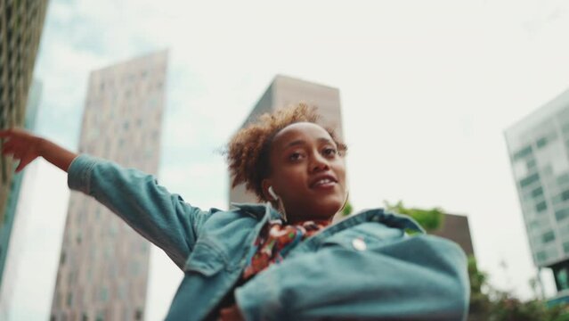 Closeup, smiling African girl with ponytail wearing denim jacket, in crop top with national pattern listening to music on headphones and dancing outdoors. Camera moving forwards approaching 
