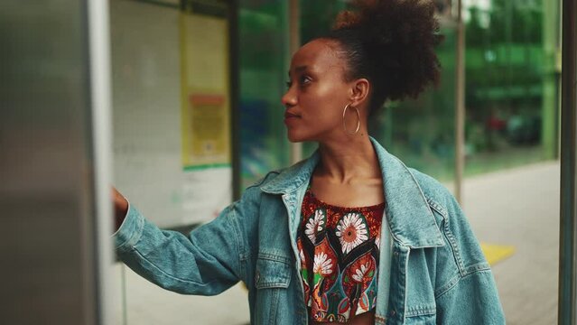 Cute African girl with ponytail, wearing denim jacket, in crop top with national pattern, standing at the bus stop and looking at the bus schedule. Slow motion