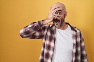 Hispanic man with beard standing over yellow background peeking in shock covering face and eyes...