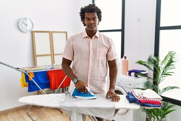 African man with curly hair ironing clothes at home relaxed with serious expression on face. simple...