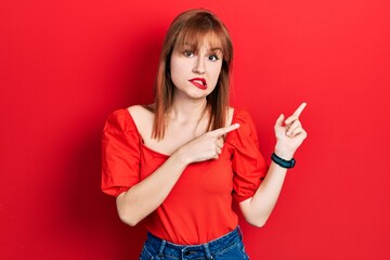 Redhead young woman wearing casual red t shirt pointing aside worried and nervous with both hands, concerned and surprised expression