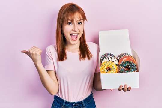 Redhead young woman holding tasty colorful doughnuts pointing thumb up to the side smiling happy with open mouth