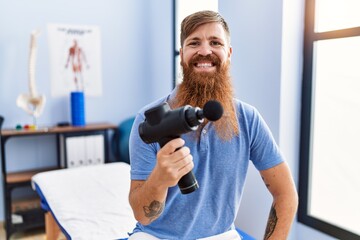 Young redhead man wearing physiotherapist uniform holding percussion massage pistol at physiotherapy clinic