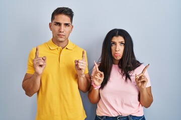 Young couple standing over isolated background pointing up looking sad and upset, indicating direction with fingers, unhappy and depressed.