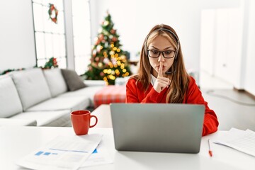 Young caucasian girl sitting on the table working using laptop by christmas tree asking to be quiet...