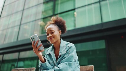 Cute African girl with ponytail, wearing denim jacket, in crop top with national pattern, sitting on bench and using smartphone on modern buildings background. Slow motion