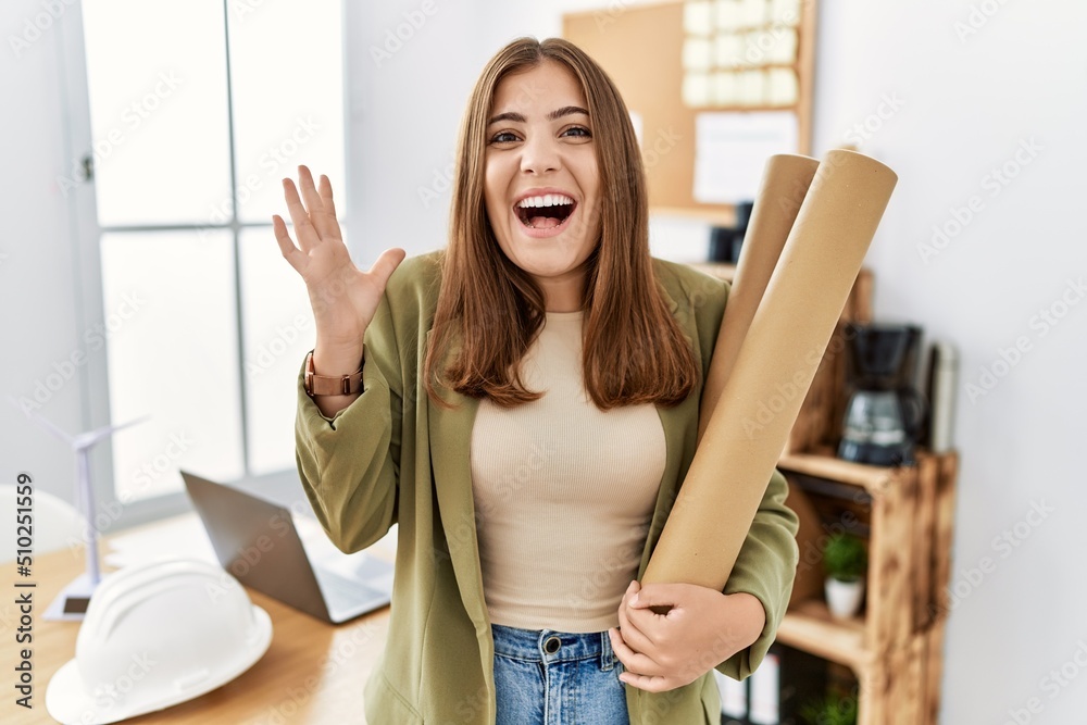 Sticker young brunette woman holding paper blueprints at the office celebrating victory with happy smile and