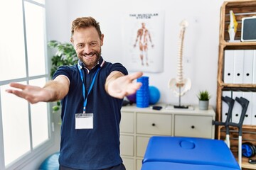Fototapeta na wymiar Middle age physiotherapist man working at pain recovery clinic looking at the camera smiling with open arms for hug. cheerful expression embracing happiness.