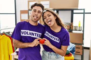 Young hispanic couple wearing volunteer t shirt holding heart sticking tongue out happy with funny expression.