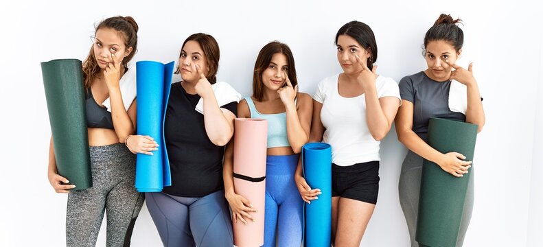 Group of women holding yoga mat standing over isolated background pointing to the eye watching you gesture, suspicious expression
