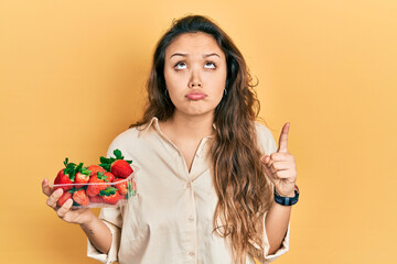Young hispanic girl holding strawberries pointing up looking sad and upset, indicating direction...