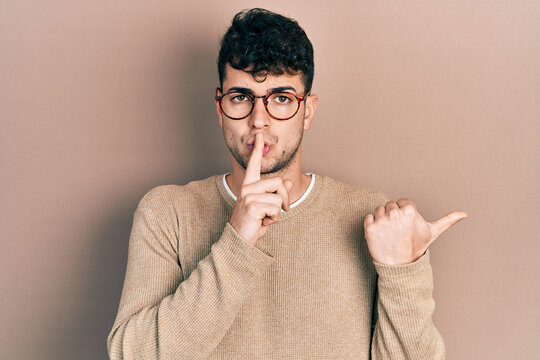 Young hispanic man wearing casual clothes and glasses asking to be quiet with finger on lips pointing with hand to the side. silence and secret concept.