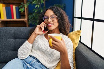 Young latin woman drinking coffee and talking on the smartphone at home