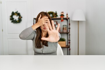 Young latin woman sitting on the table by christmas decor covering eyes with hands and doing stop gesture with sad and fear expression. embarrassed and negative concept.