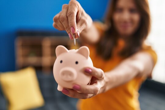 Young latin woman smiling confident insert coin on piggy bank at home