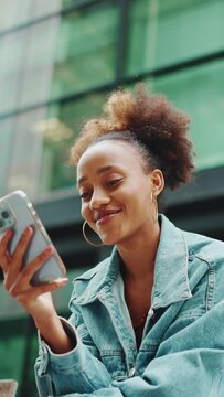 VERTICAL VIDEO:Closeup, cute African girl with ponytail, wearing denim jacket, in crop top with national pattern, sitting at the bus stop and using her mobile phone. Slow motion