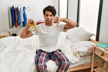 Young hispanic man holding alarm clock sitting on the bedroom with angry face, negative sign...