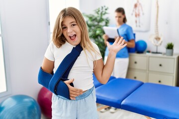 Blonde little girl wearing arm on sling at rehabilitation clinic celebrating victory with happy smile and winner expression with raised hands