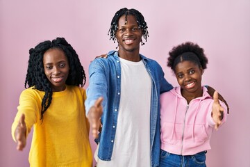 Group of three young black people standing together over pink background smiling friendly offering...