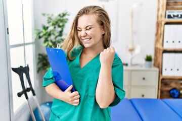 Young caucasian woman working at pain recovery clinic very happy and excited doing winner gesture with arms raised, smiling and screaming for success. celebration concept.