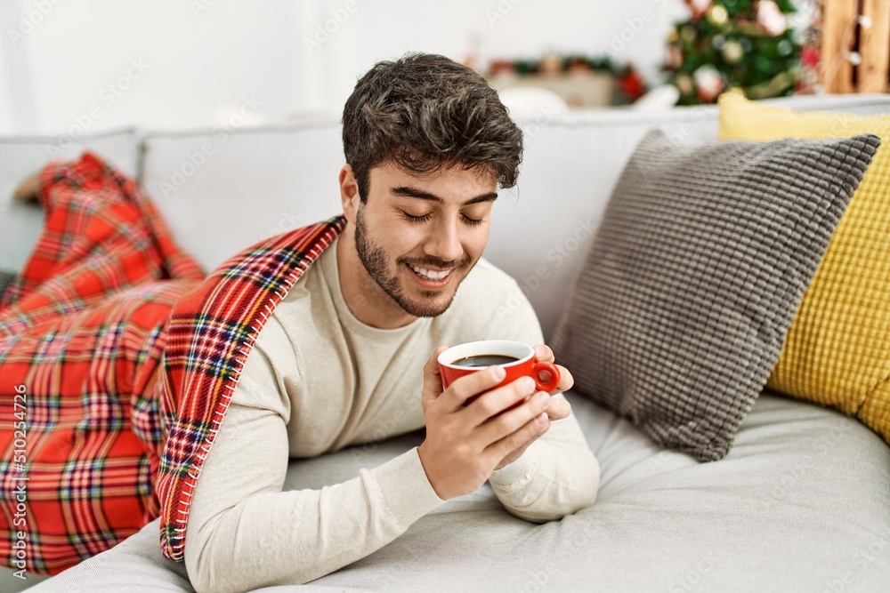 Poster Young hispanic man smiling happy lying on the sofa drinking coffee at home.