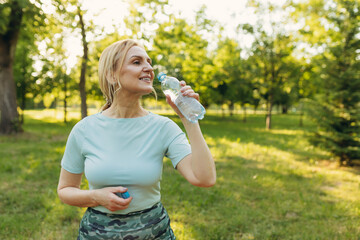 Mature woman drinks water while exercising in the park. The concept of a healthy lifestyle. Copy space