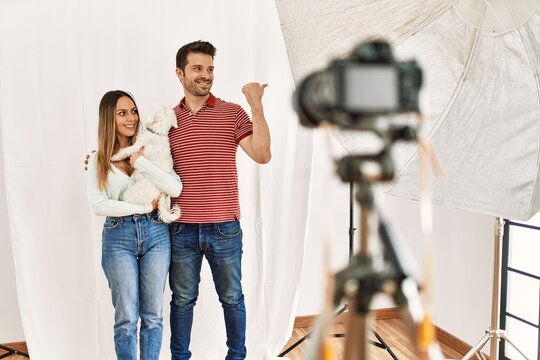 Couple of boyfriend and girlfriend with dog posing as model at photography studio pointing thumb up to the side smiling happy with open mouth