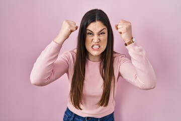 Young brunette woman standing over pink background angry and mad raising fists frustrated and...