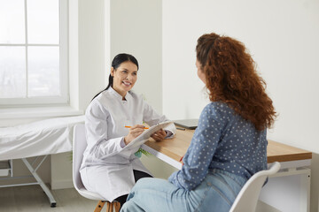 Doctor at modern clinic or medical office giving consultation to patient. Female physician talking to young woman, asking questions, listening carefully, holding clipboard and writing down symptoms