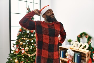 African american man wearing santa claus hat standing by christmas tree stretching back, tired and...