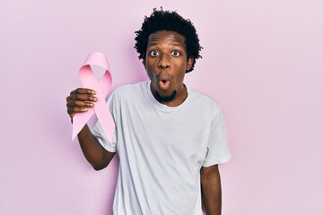 Young african american man holding pink cancer ribbon scared and amazed with open mouth for surprise, disbelief face