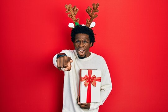 Young african american man wearing deer christmas hat holding gift pointing displeased and frustrated to the camera, angry and furious with you