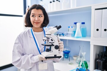 Young hispanic woman working at scientist laboratory holding microscope smiling looking to the side and staring away thinking.