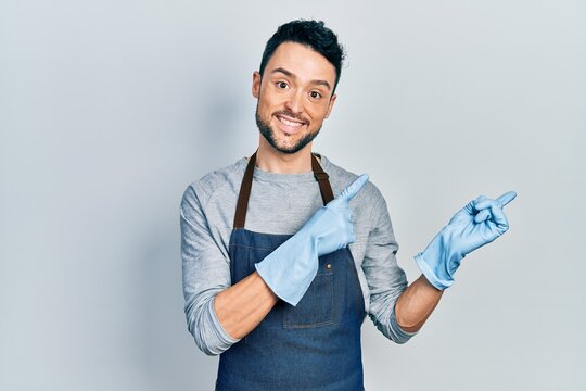 Young hispanic man wearing apron and gloves smiling and looking at the camera pointing with two hands and fingers to the side.