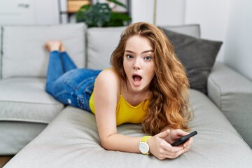 Young caucasian woman lying on the sofa using smartphone in shock face, looking skeptical and sarcastic, surprised with open mouth - Powered by Adobe