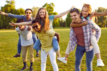 Fototapeta premium Friends gaving fun outdoors. Six cheerful young friends carry each other on their backs while walking in park. Multiracial men and women have fun together during summer walk outdoors.