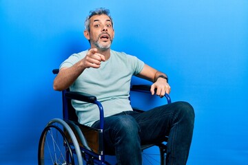 Handsome middle age man with grey hair sitting on wheelchair pointing displeased and frustrated to the camera, angry and furious with you