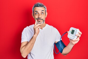 Handsome middle age man with grey hair using blood pressure monitor serious face thinking about...