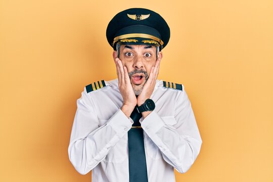 Handsome middle age man with grey hair wearing airplane pilot uniform afraid and shocked, surprise and amazed expression with hands on face