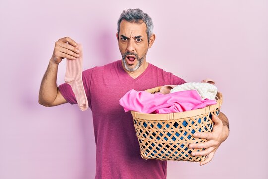 Handsome middle age man with grey hair holding laundry basket and dirty sock in shock face, looking skeptical and sarcastic, surprised with open mouth