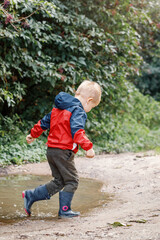 Child in a red waterproof coat in the rain jumping in puddles. A boy stands with his head bowed, a bright raincoat. Kid playing autumn park. Outdoor fun by any weather.