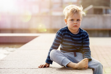 Little sad boy sitting by the fountain on a Sunny day, the child looks to photo camera, there is...